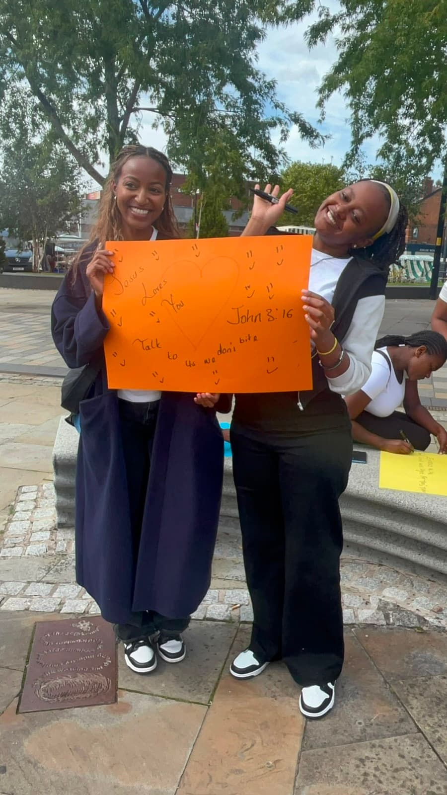 Two young women smiling while holding a sign about Jesus loving people